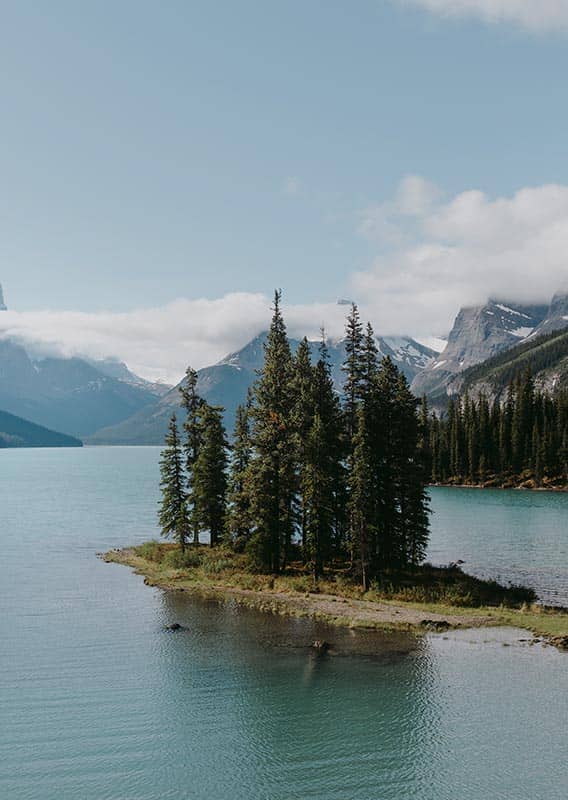 A small tree-covered peninsula on a calm lake below tall snow-covered mountains.