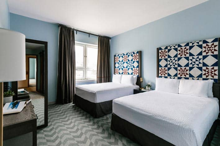 Two beds with red and blue quilted headboards, with sun shining through a window.