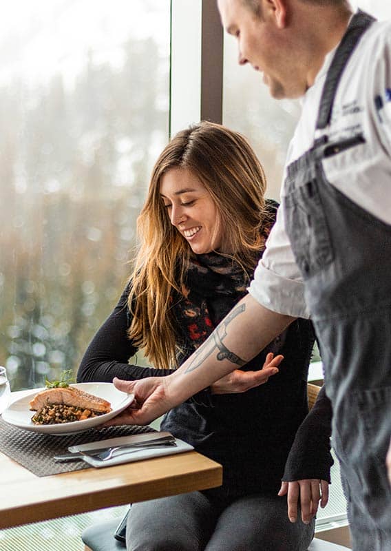 A server brings a dinner plate to a table at a windowside.