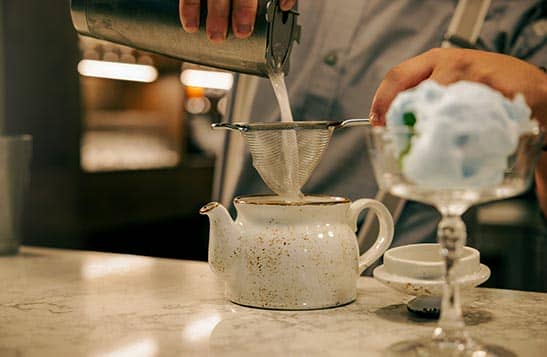 A bartender pours a drink through strainer into a ceramic glazed pot.