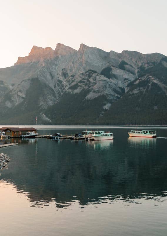 A few boats around a dock extending out on a calm lake below rocky mountains.