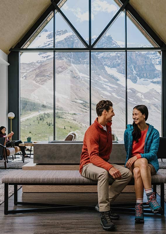 Two people sit on a bench in a lounge with a high ceiling and tall window.