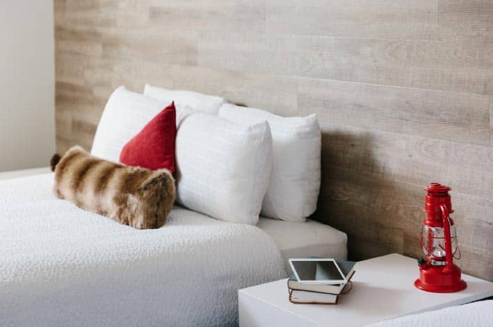 A bed against a rustic wooden wall and bedside table with books and a red lamp.