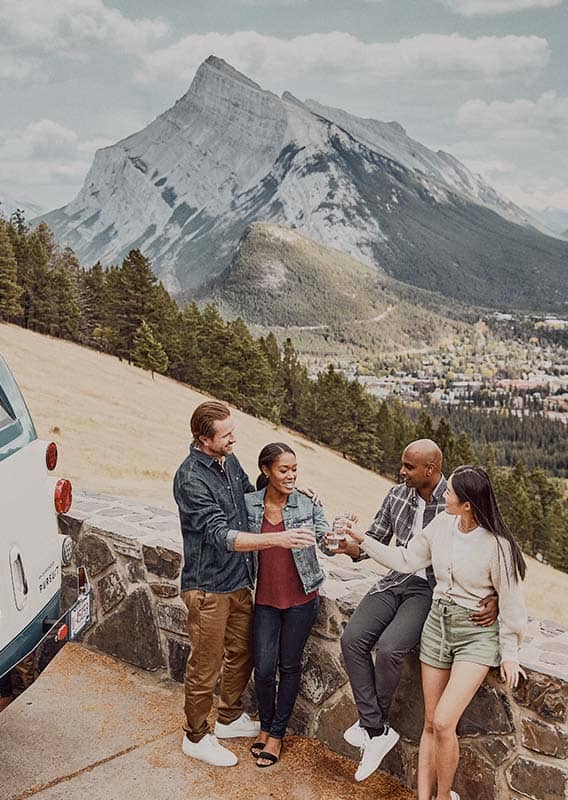 A group of people stand at a stone wall, overlooking a grassy hillside and town below mountains.