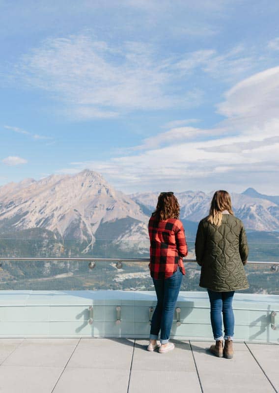 Two people stand at a viewpoint overlooking a wide valley between mountains.