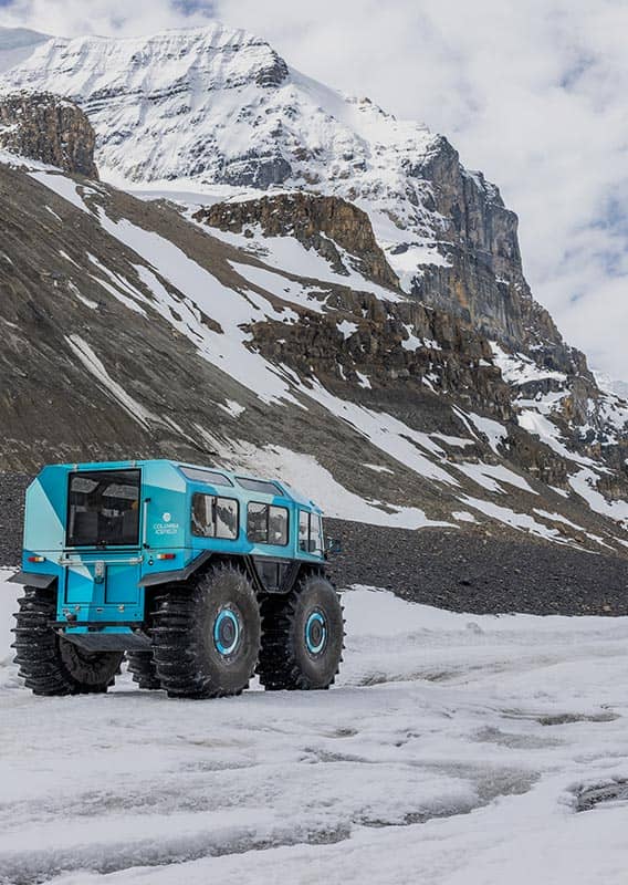 An all terrain Sherp vehicle driving on a glacier below tall mountains.