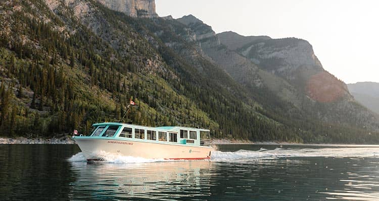 A blue and white boat cruises on a lake below a forested mountainside.