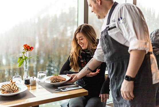 A chef brings a dinner plate to a diner at a windowside seat.