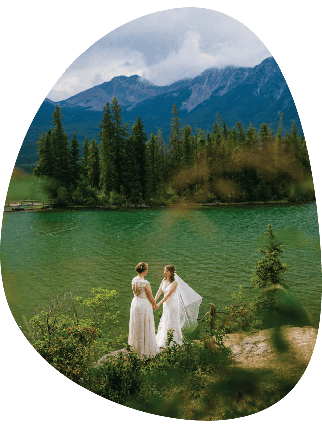 Two brides hold hands at a rocky viewpoint above a small mountain lake.