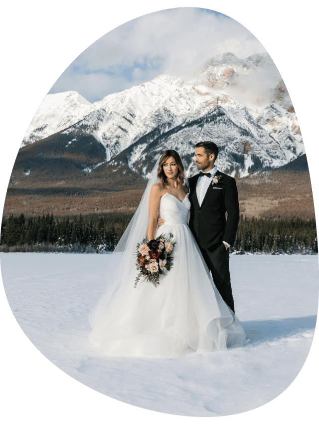 Two brides hold hands at a rocky viewpoint above a small mountain lake.